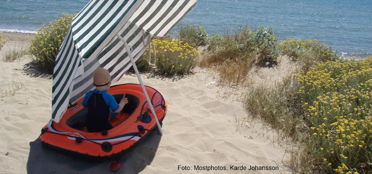 Bilden visar ett barn som sitter i en gummibåt på stranden, i skydd av ett parasoll.