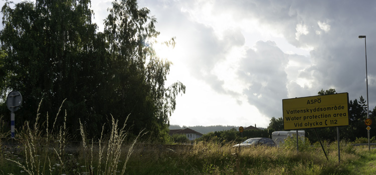 Bilden visar en skylt som visar var Aspö vattenskyddsområde börjar. Bakom skylten kommer en bil med släpvagn.