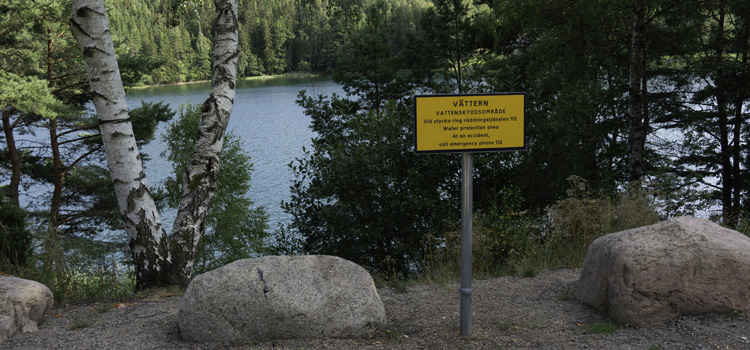 En skylt som visar att här är du inom Vätterns vattenskyddsområde. Bakom skylten finns träd i en slänt ner mot vattnet.