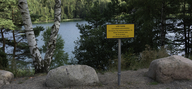 En skylt som visar att här är du inom Vätterns vattenskyddsområde. Bakom skylten finns träd i en slänt ner mot vattnet.